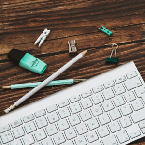 bottom-square-flatlay-blue-gold-teal-stationary-scattered-across-bottom-image-with-mac-keyboard-gold-clear-plant-pot-across-a-brown-wooden-desk