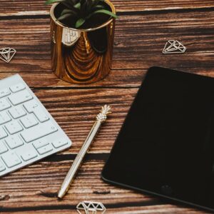 iPad-in-corner-of-right-hand-screen-gold-pineapple-pen-macbook-keyboard-on-left-gold-plant-pot-green-spikey-leaves-gold-stationary-on-a-brown-wooden-desk