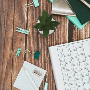 square-flatlay-blue-gold-teal-stationary-scattered-across-side-image-with-mac-keyboard-in-bottom-right-corner-gold-blue-teal-notepads-clear-plant-plot-across-a-brown-wooden-desk