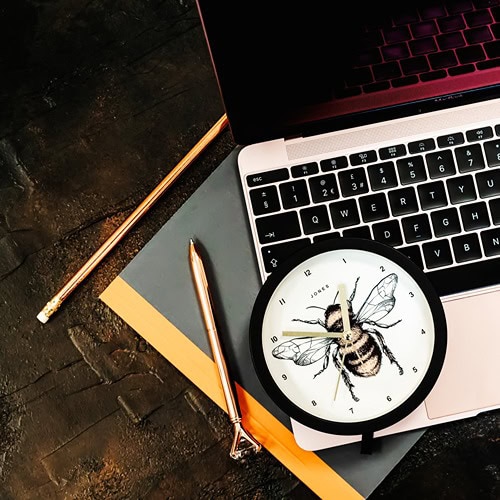 black-circle-framed-desk-clock-with-coloured-bee-drawing-laid-on-open-laptop-keys-with-charcoal-notebook-rose-gold-pen-and-pencil-on-bronze-textured-surface